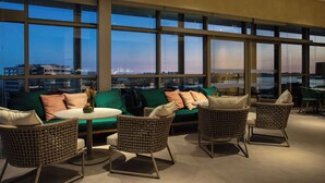 A modern lounge area with a green sofa, wicker chairs, and a round table. Large windows offer a view of the city skyline. - Hilton Barra Rio De Janeiro (Rio de Janeiro)