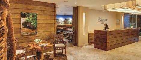 Un espace d’accueil avec un comptoir de réception en bois, un coin salon avec des chaises et une table, et un tableau représentant un paysage accroché au mur.