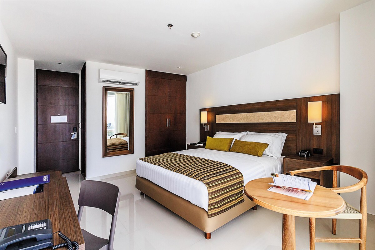 Quarto de hotel com cama, mesa de cabeceira, cadeira, escrivaninha e abajur.
