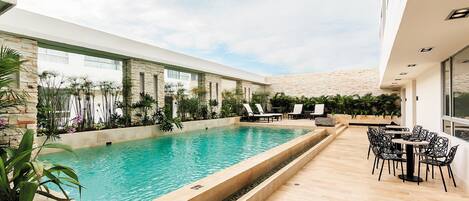 A modern indoor pool area with a clear view of the sky, surrounded by lounge chairs and tables, and a stone wall in the background.