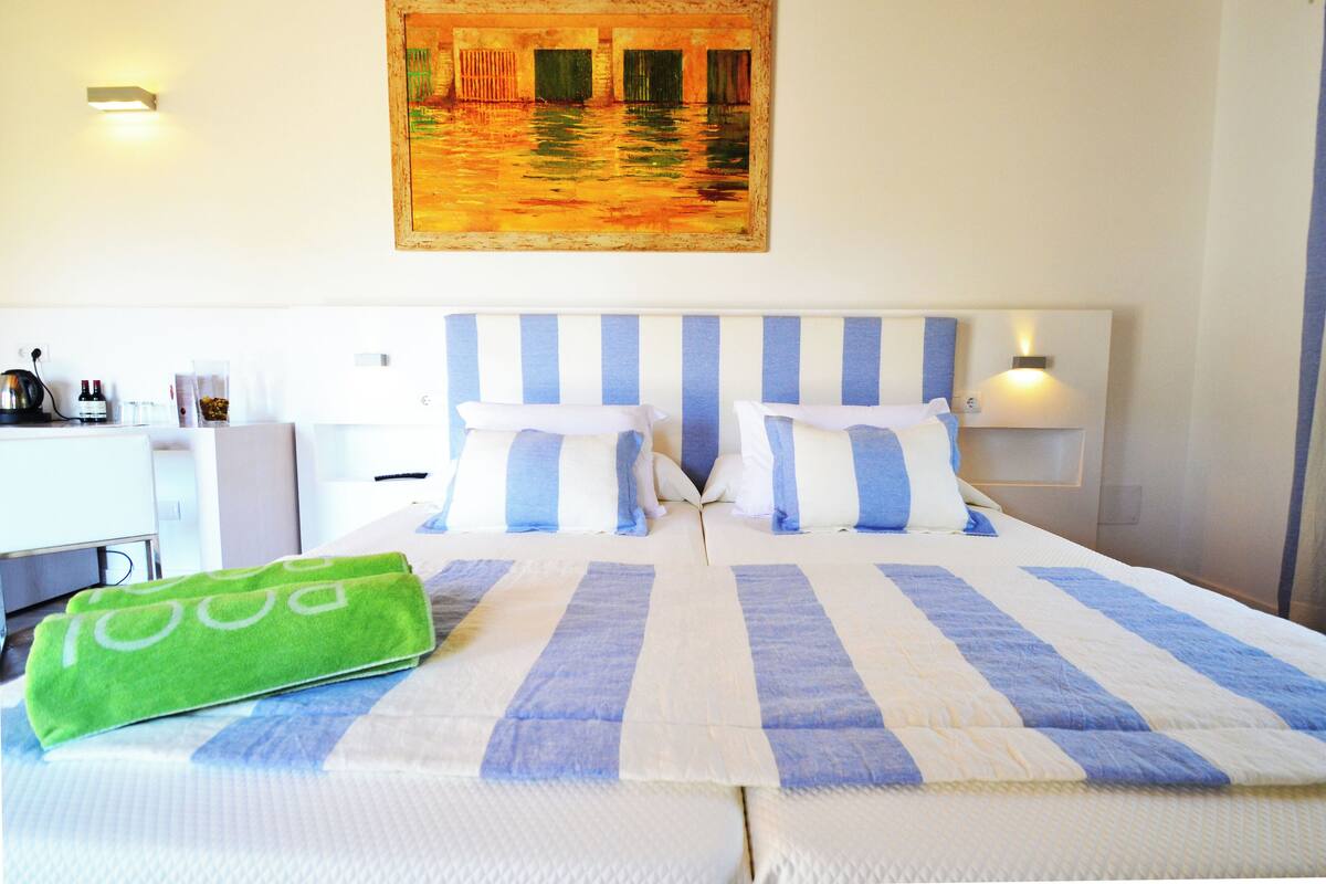 A hotel room with a bed, pillows, a green towel, a painting, and a desk with a lamp.