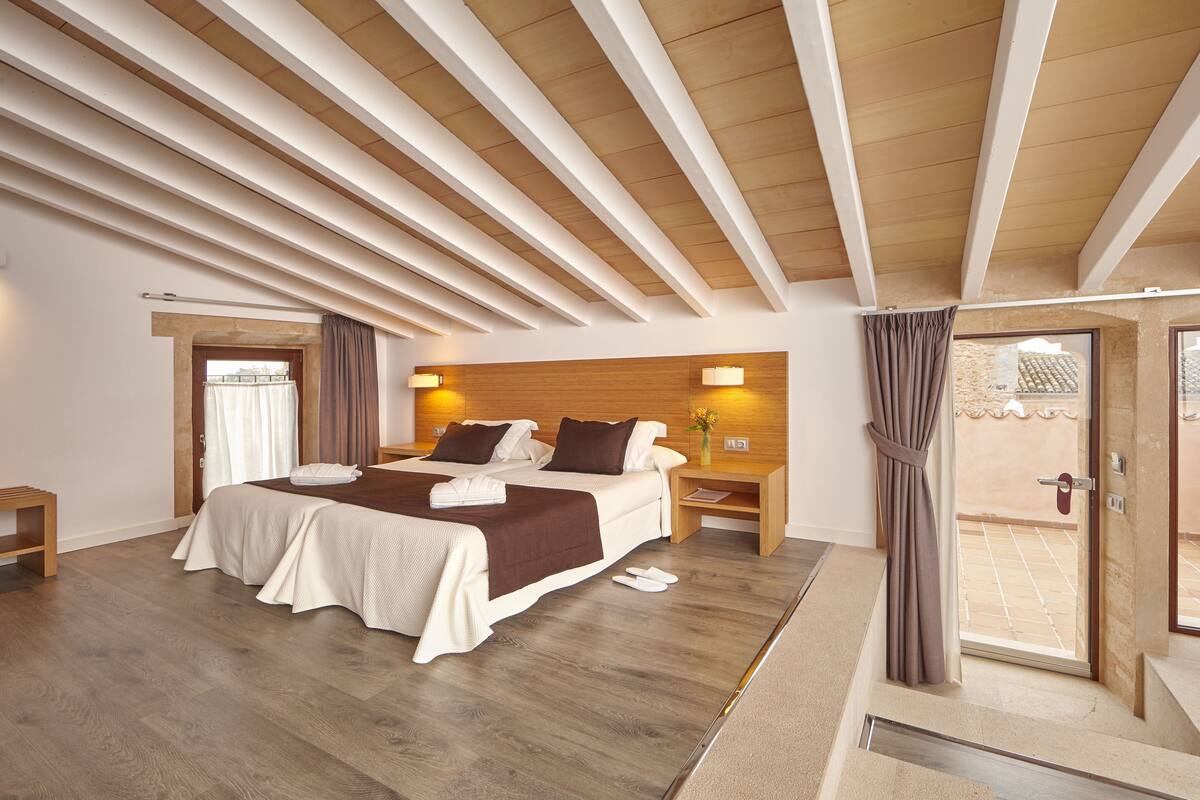 A modern hotel room with a large bed, wooden ceiling beams, a small bedside table, and a view of the outside balcony.