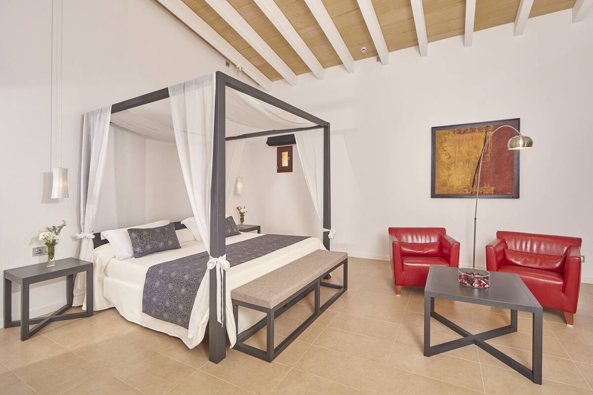 A four-poster bed with a bench, two red leather chairs, a small table, and a painting on the wall.