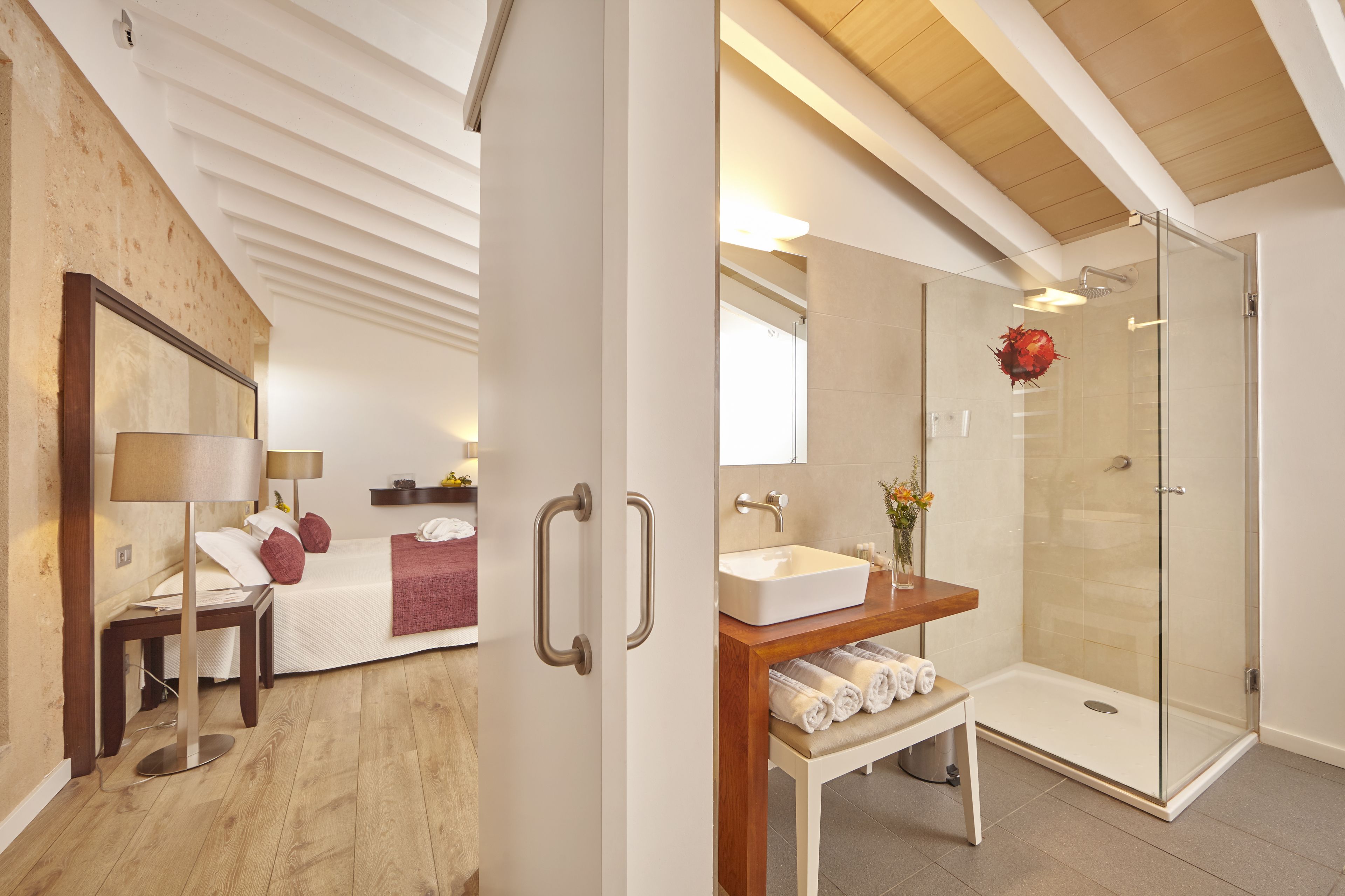 Une chambre moderne comprenant un lit, une table de chevet, une lampe et un grand miroir. À côté se trouve une salle de bain avec une douche en verre, un lavabo et un porte-serviettes.