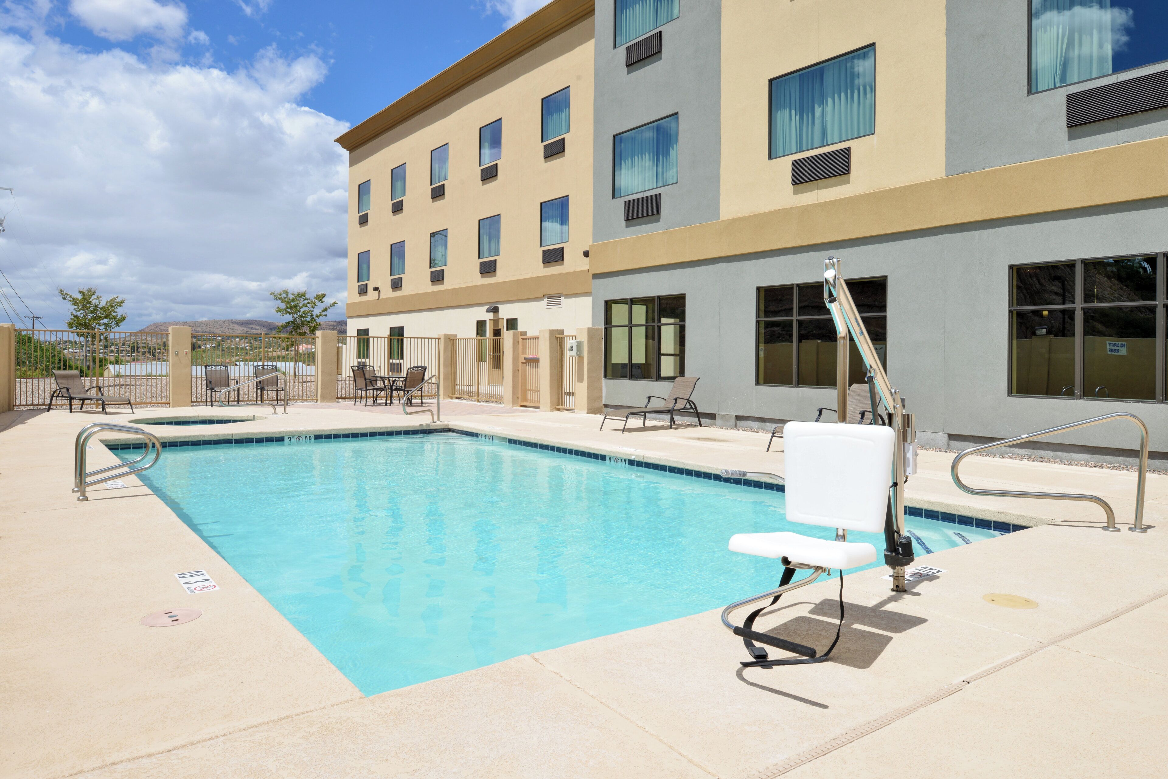 A swimming pool with a lifeguard chair and a sun chair, surrounded by a fence and a building with multiple windows.
