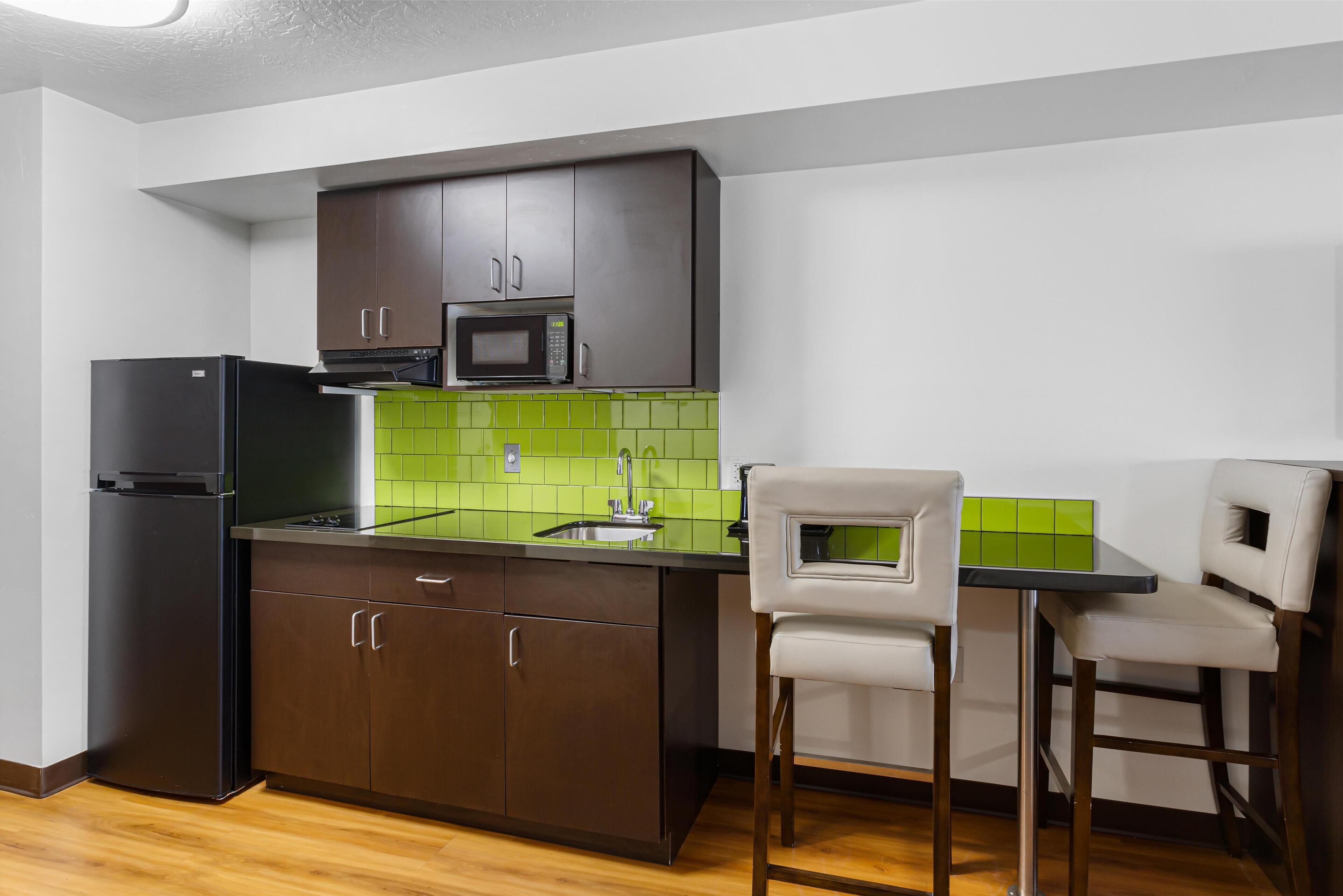 A compact kitchen with dark wooden cabinets, a black refrigerator, a microwave, and a green tiled backsplash. There is a small bar area with two chairs.