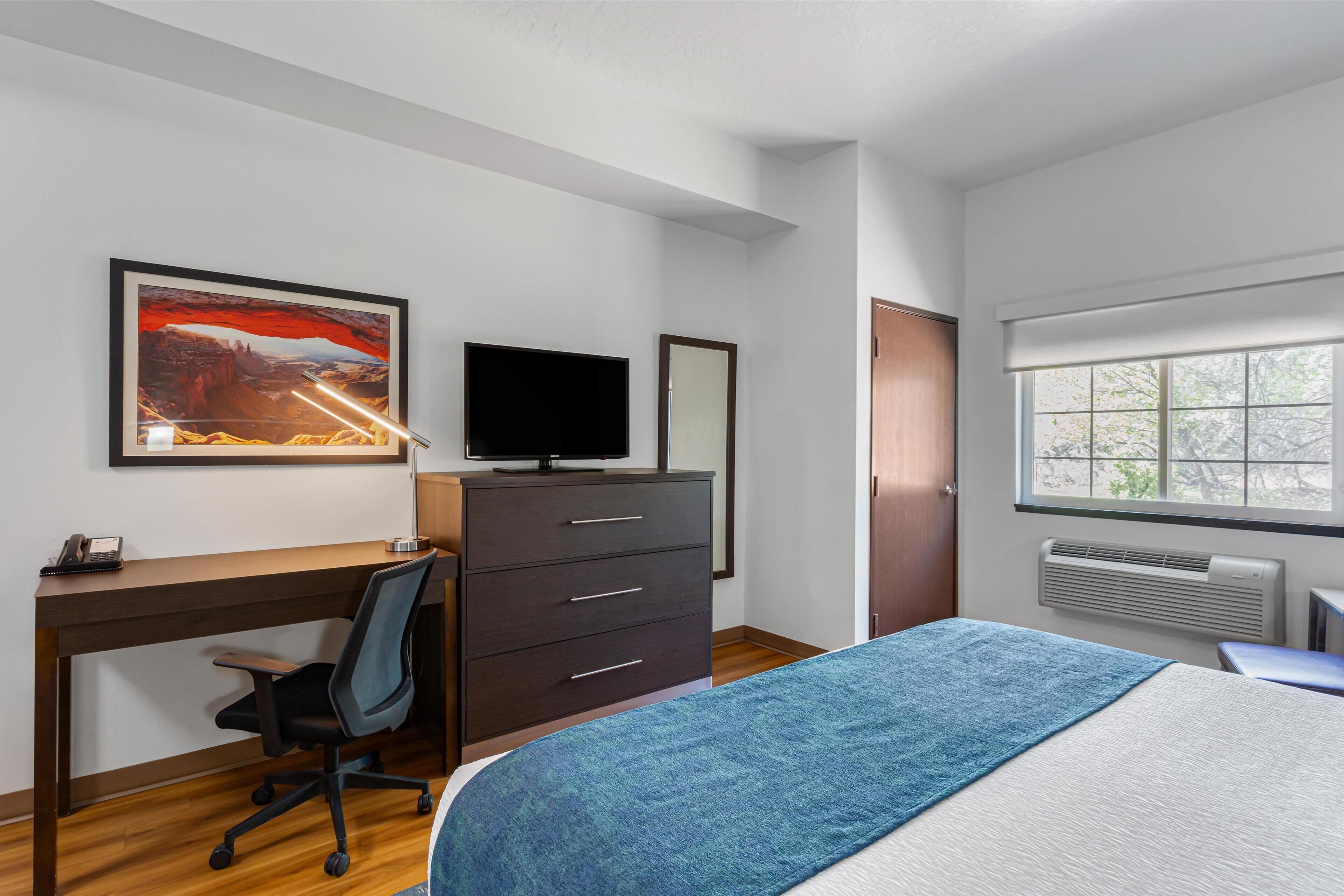 Une chambre moderne avec un bureau en bois, une télévision, un lit recouvert d’une couverture bleue et une fenêtre donnant sur des arbres.