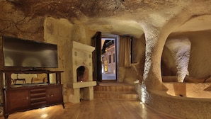 A cave-like interior with a television on a wooden cabinet, stone walls, and a staircase leading to an upper level. - Golden Cave Suites (Ürgüp)