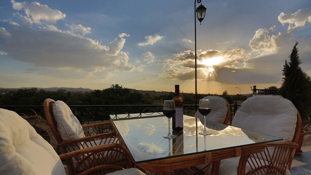 A glass table with two chairs and a bottle of wine, overlooking a scenic landscape with mountains and trees. - Golden Cave Suites (Ürgüp)