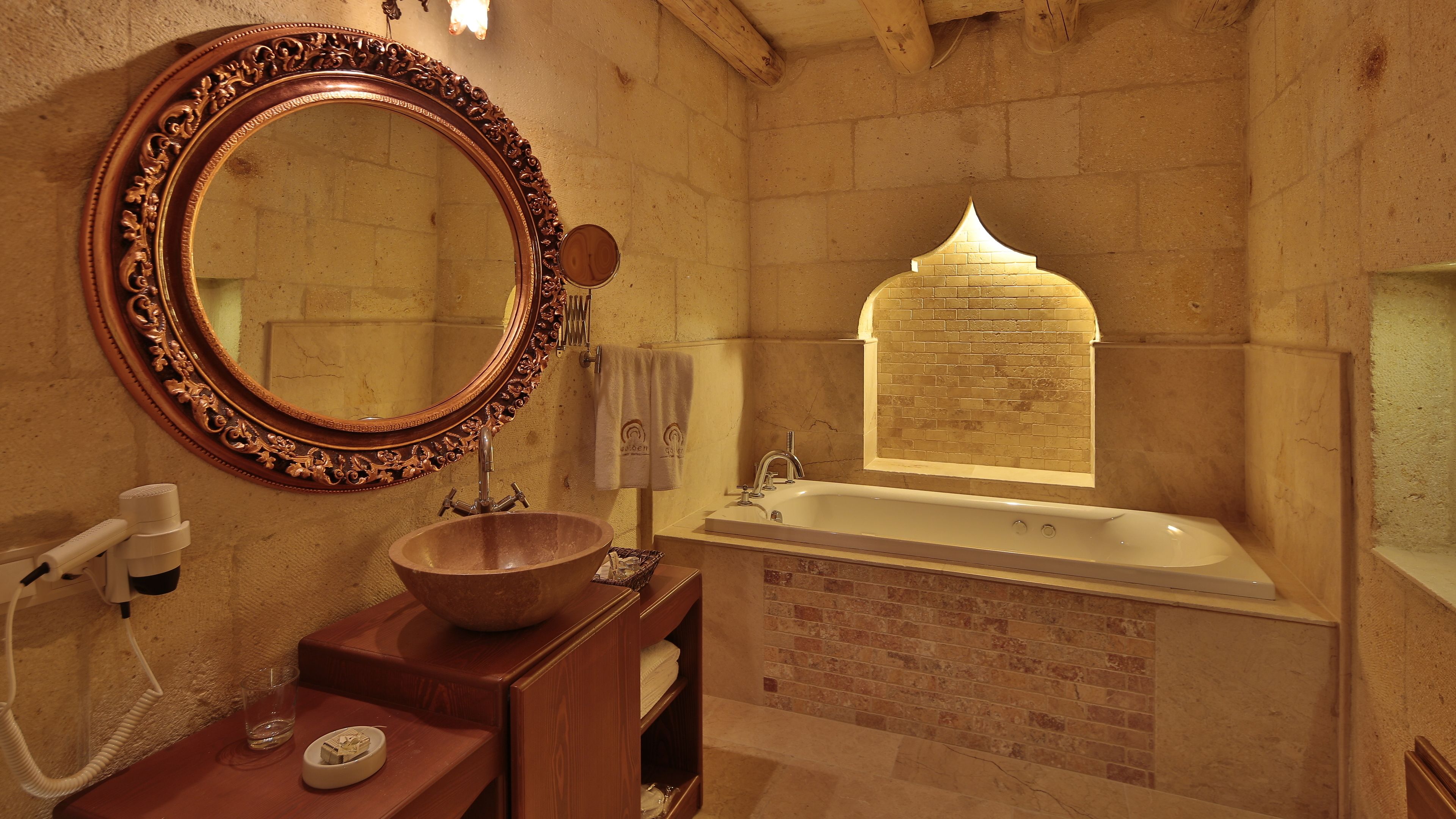 A bathroom with a wooden vanity, a large ornate mirror, a freestanding bathtub, and a window with a brick pattern.