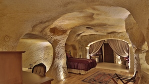 A room with a rustic stone ceiling, a wooden desk, a bed with a purple bedspread, and a patterned rug. - Golden Cave Suites (Ürgüp)