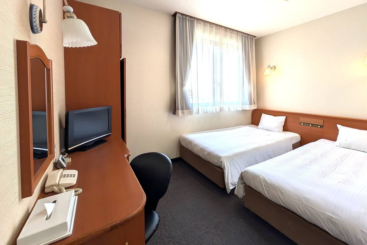 Ein Hotelzimmer mit zwei Betten, einem Schreibtisch, einem Fernseher, einem Telefon und einem Fenster mit Vorhängen.