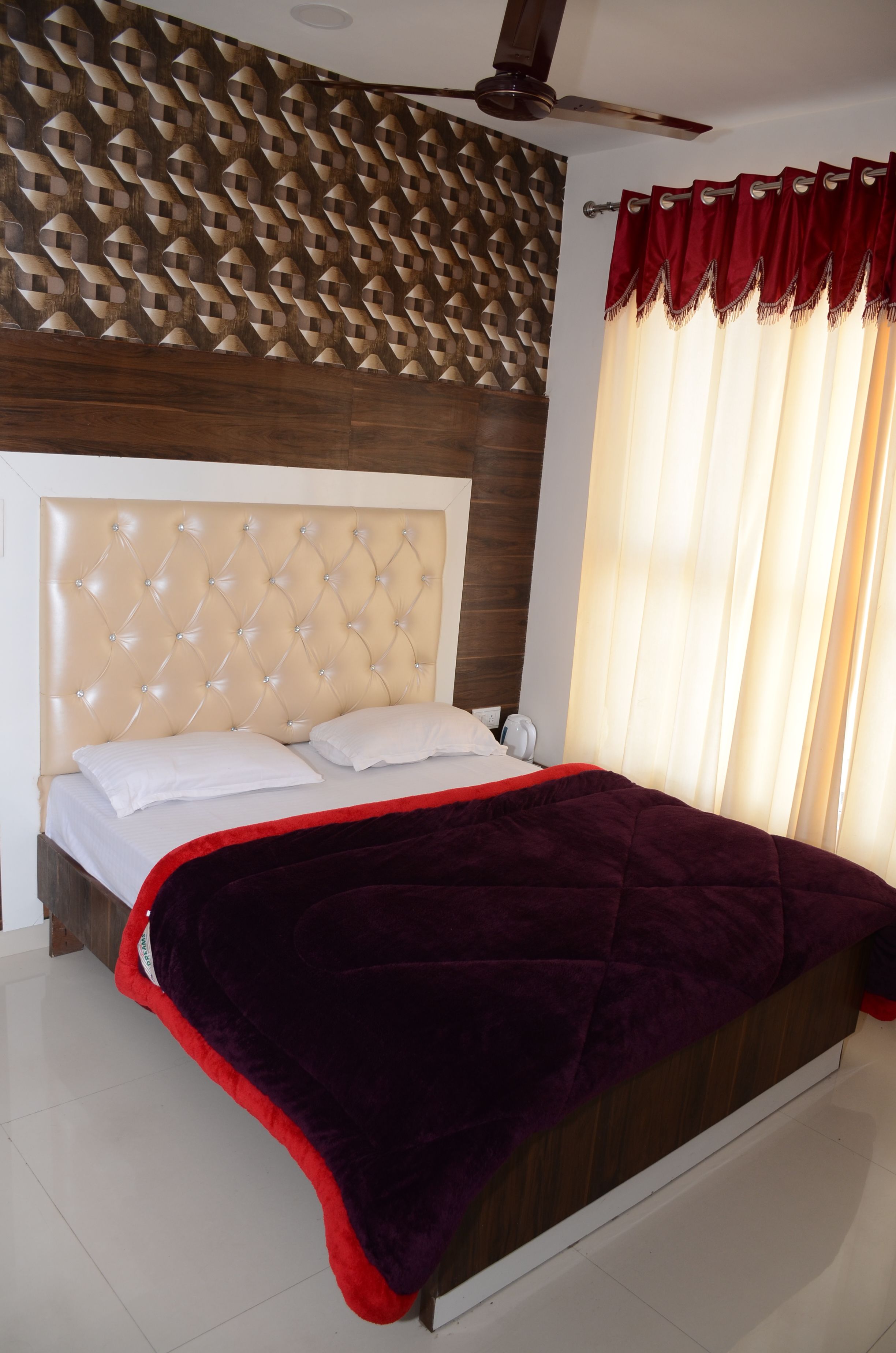 A hotel room with a tufted headboard, a red and black bedspread, a patterned wall, and a ceiling fan.