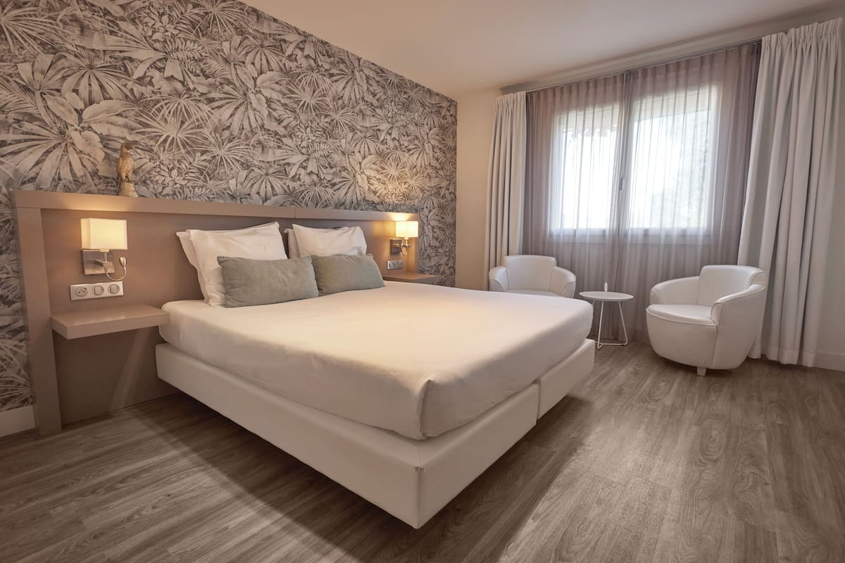 Hotellihuone, jossa on suuri sänky, kaksi yöpöytävalaisinta, pieni pöytä, tuoli ja kukallinen tapetti.