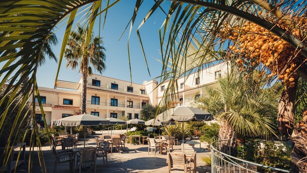 An outdoor patio area with wooden tables and chairs, umbrellas, and palm trees. - Van der Valk Hotel Le Catalogne Saint Aygulf (Fréjus)