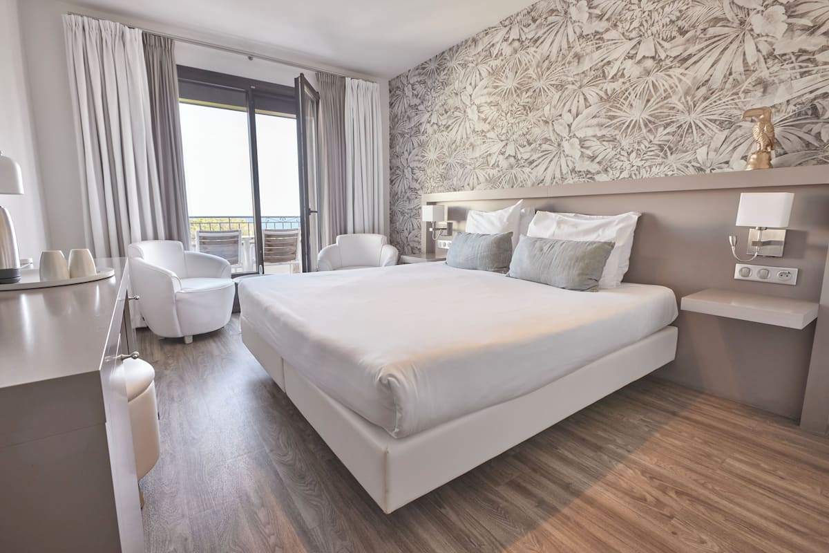 Moderni hotellihuone, jossa on suuri sänky, kaksi valkoista tuolia, työpöytä ja parveke, jossa on verhot.