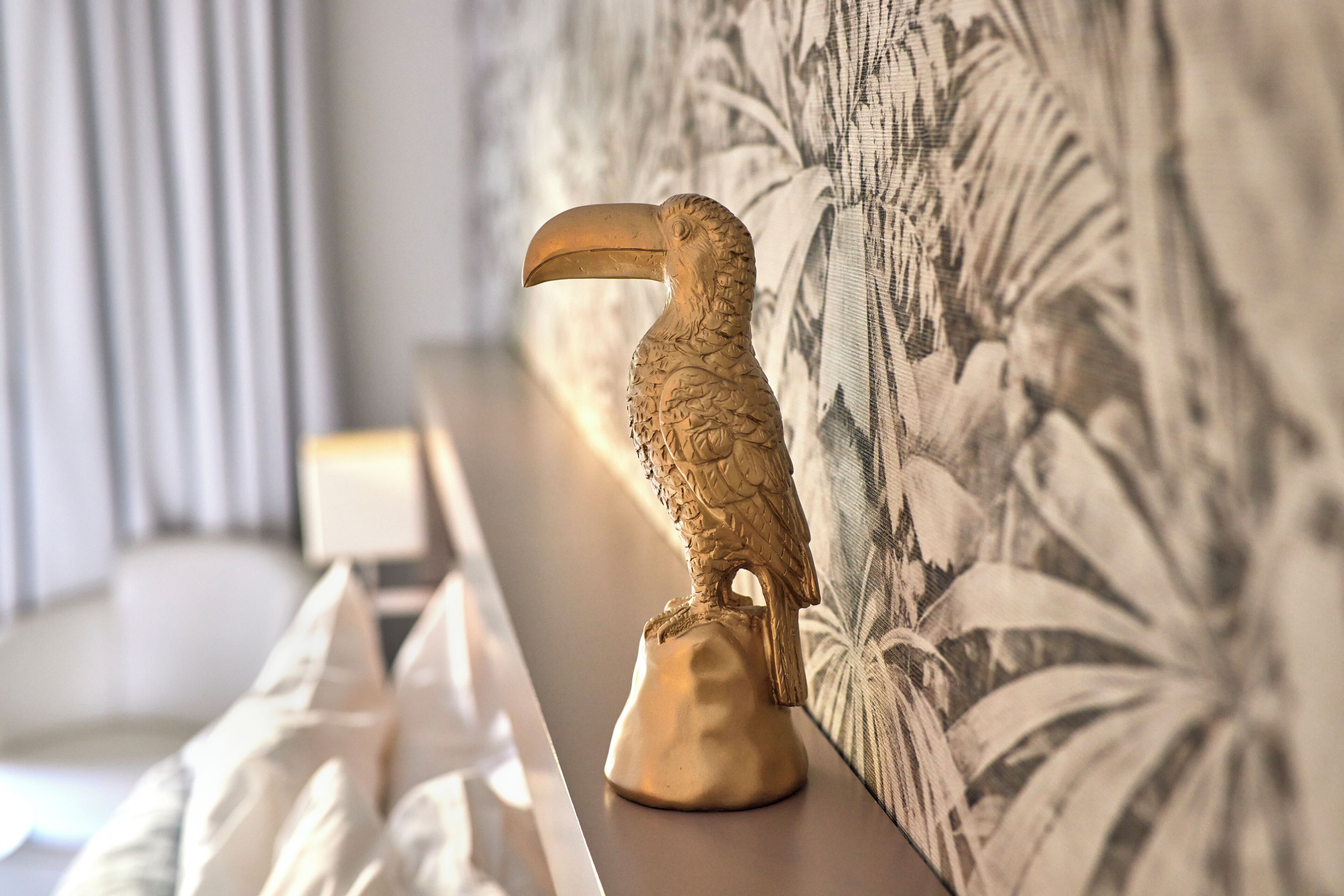 A golden toucan statue on a pedestal against a wall with a floral pattern.