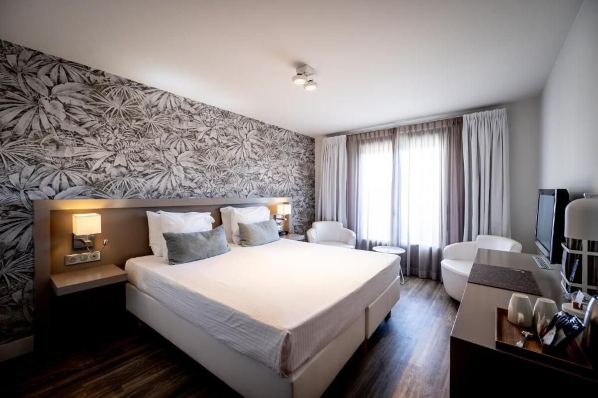 Ein Hotelzimmer mit einem großen Bett, zwei Nachttischen mit Lampen, einem Schreibtisch mit Stuhl, einem Fernseher und einem Fenster mit Vorhängen.