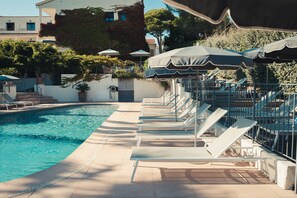A poolside area with lounge chairs and umbrellas, a clear pool, and a building with a white facade and greenery. - Van der Valk Hotel Le Catalogne Saint Aygulf (Fréjus)