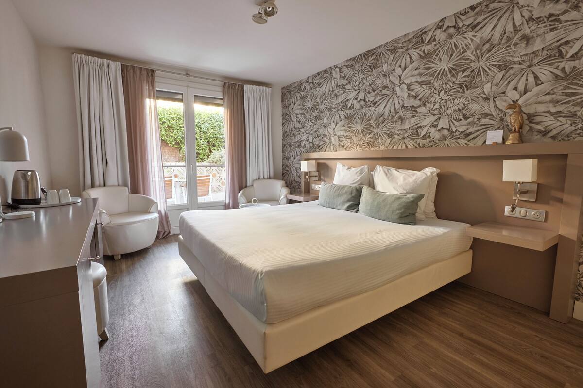 Hotellihuone, jossa on sänky, työpöytä, tuoli, ikkuna verhoilla ja trooppinen tapetti.