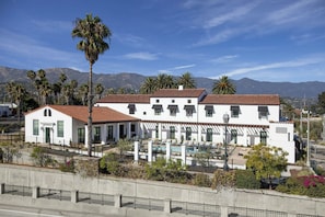A white building with a pool and palm trees, surrounded by mountains.