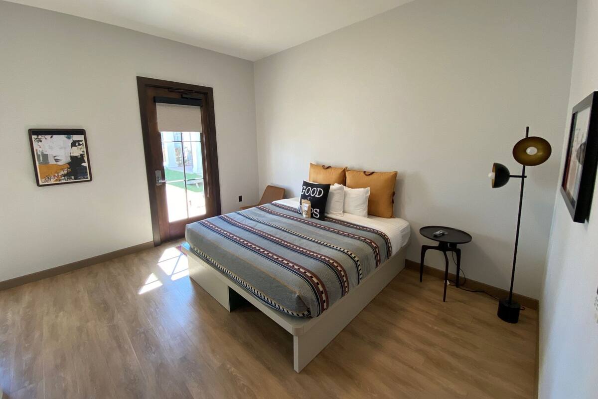 A bedroom with a bed, a small table, a floor lamp, and a door leading to a balcony.