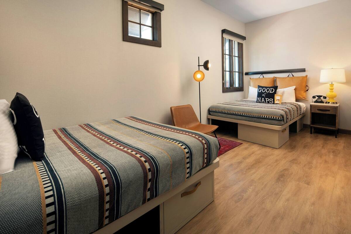 A hotel room with two beds, a nightstand with a lamp, and a window with blinds.