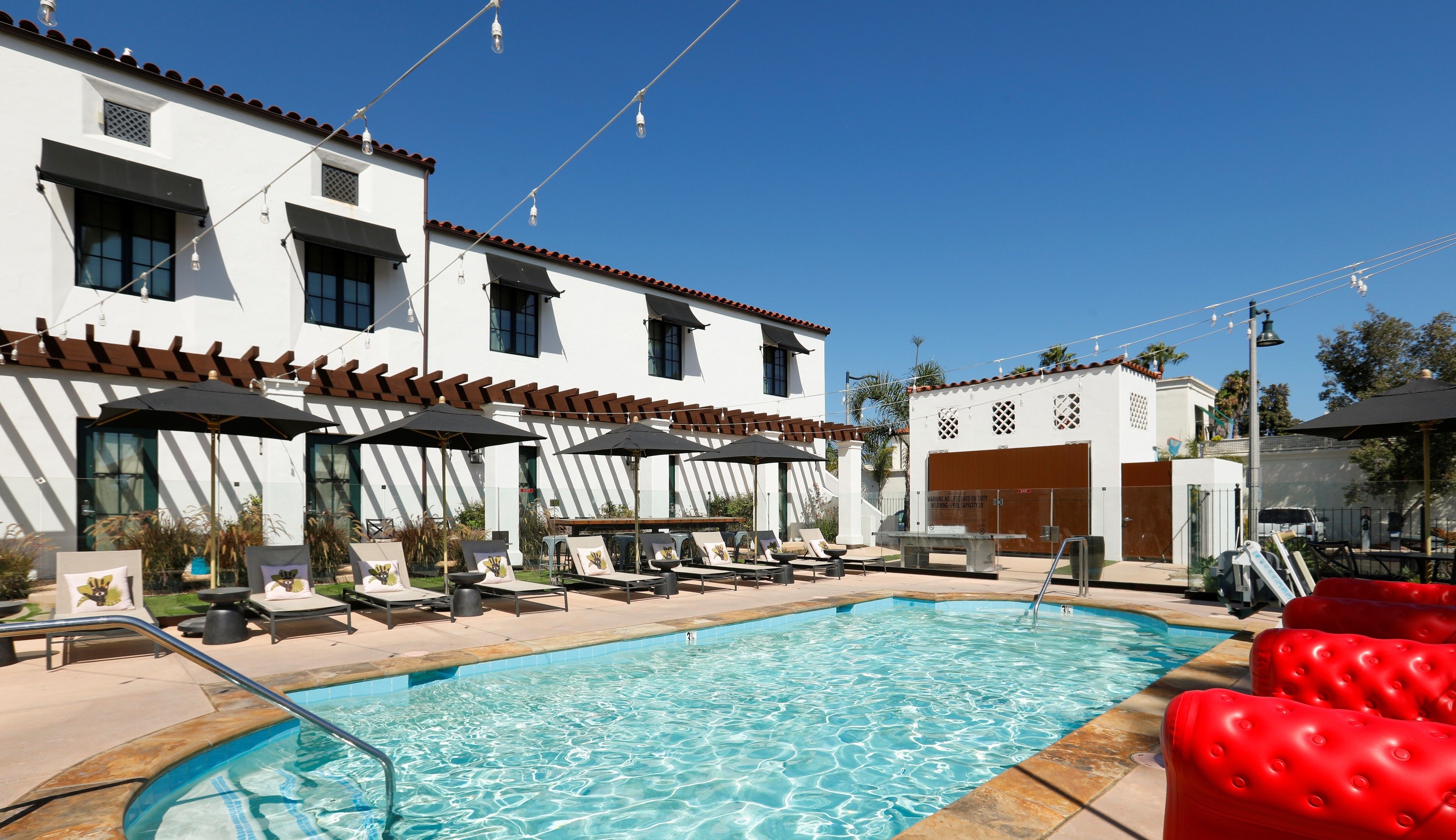 A pool area with lounge chairs and umbrellas, a building with a tiled roof, and a clear blue sky.