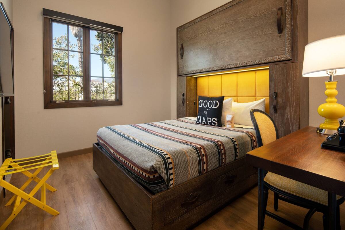 A bedroom with a bed, bedside table, lamp, and a window with a view.