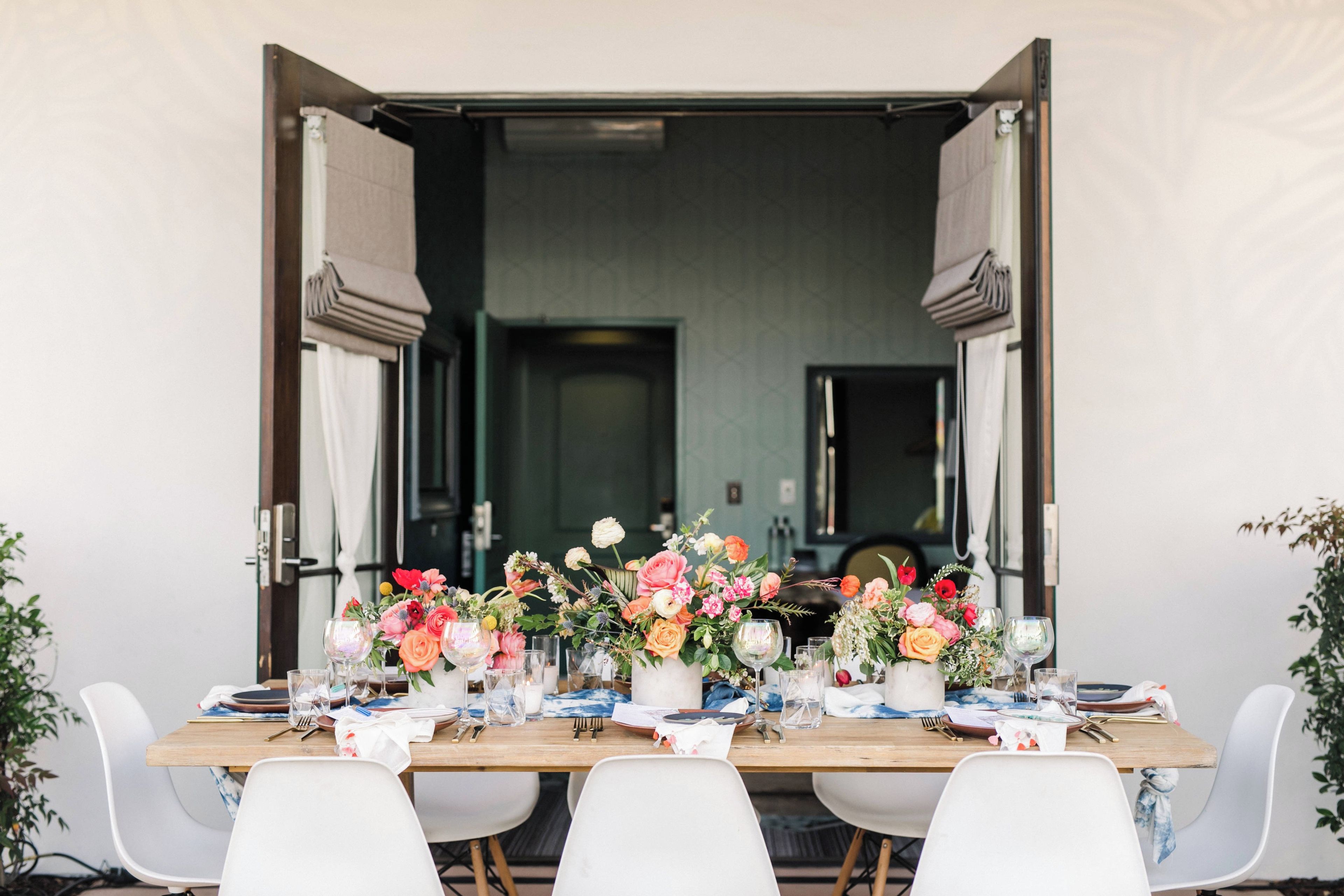Mesa de jantar posta para refeição, com cadeiras em cada lado, um arranjo floral no centro e vista para um quarto com porta e espelho.