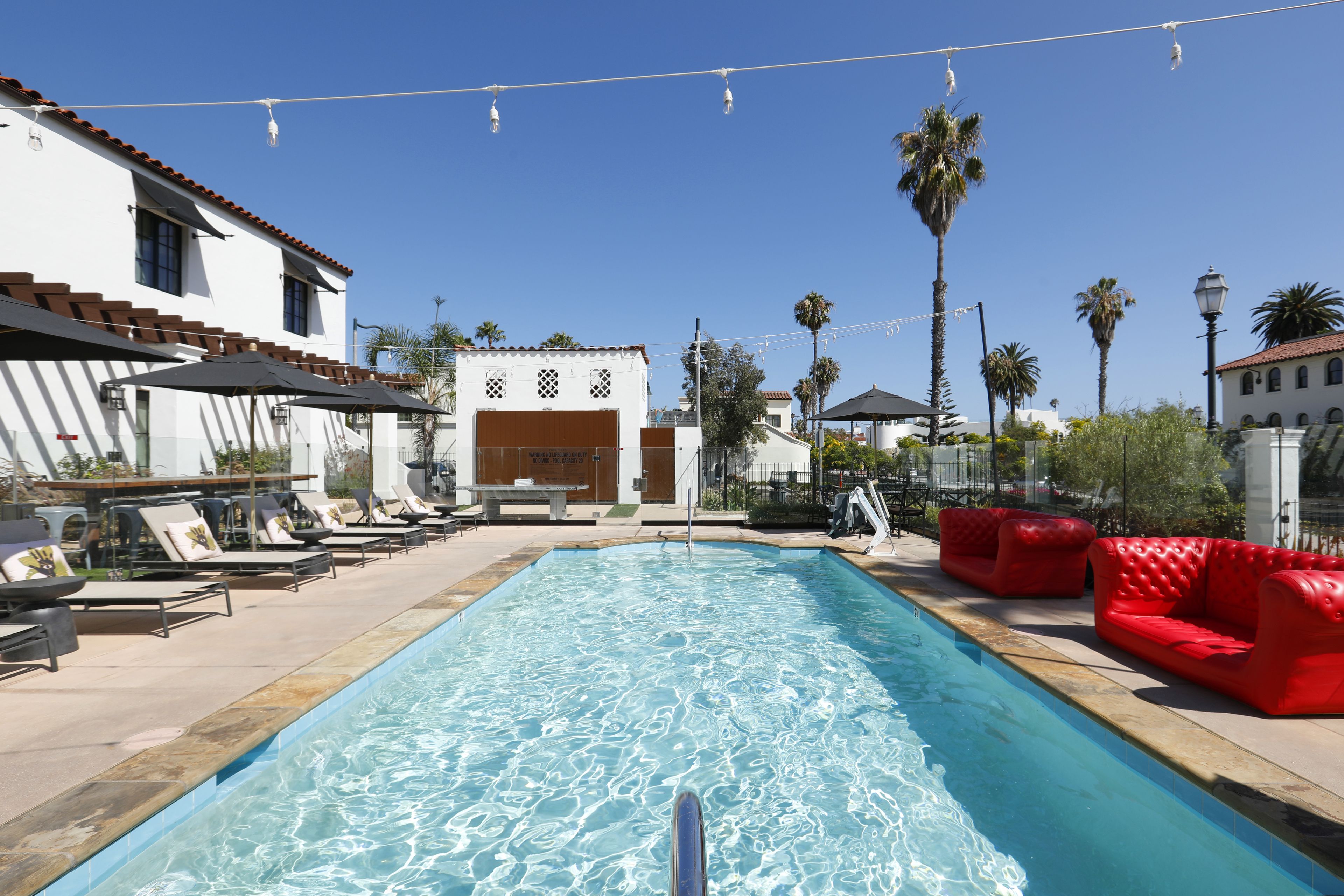 Ein Swimmingpool mit Liegestühlen und roten Sofas, umgeben von Palmen und Gebäuden.