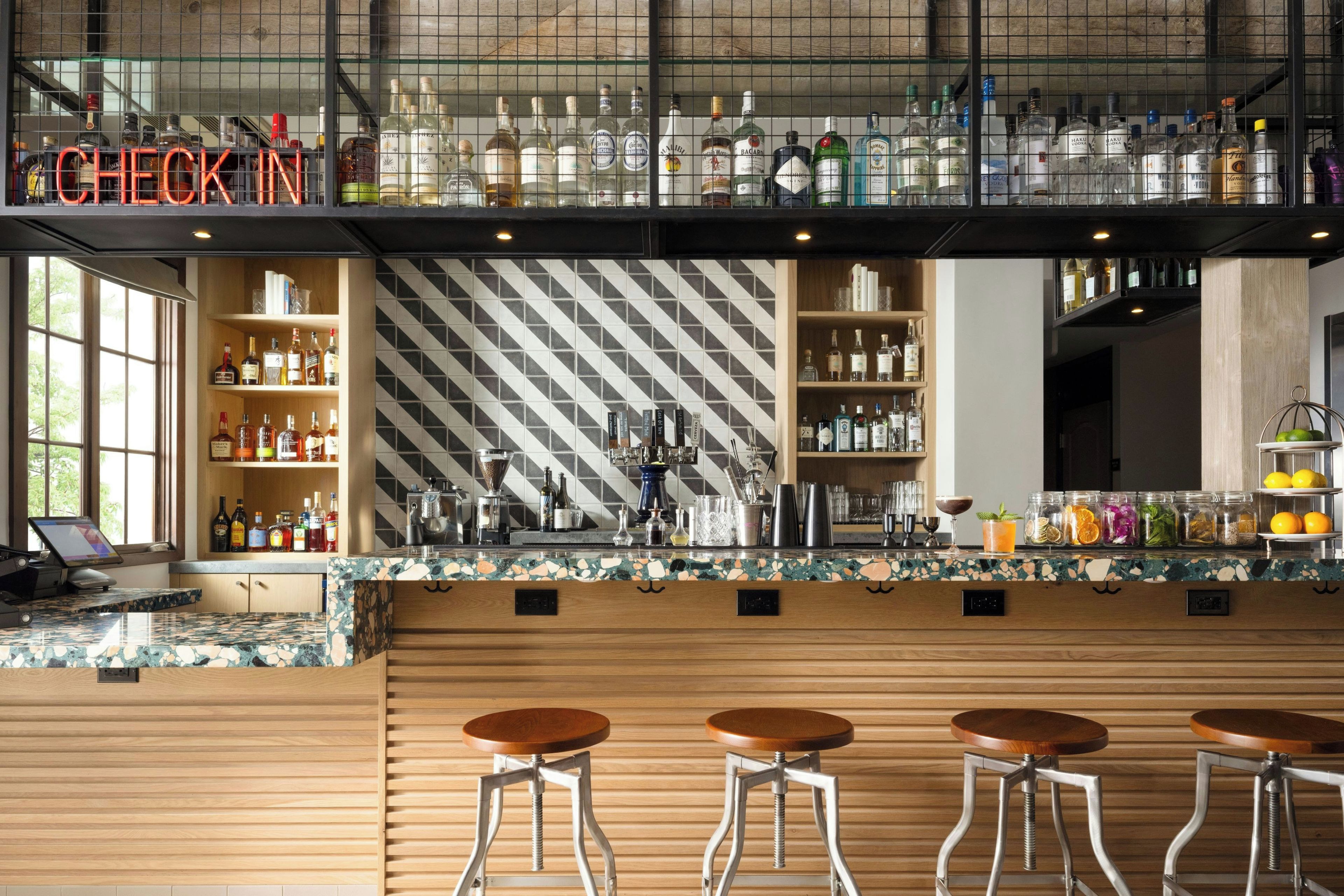 A bar area with a check-in sign, shelves of liquor bottles, a coffee machine, and bar stools.