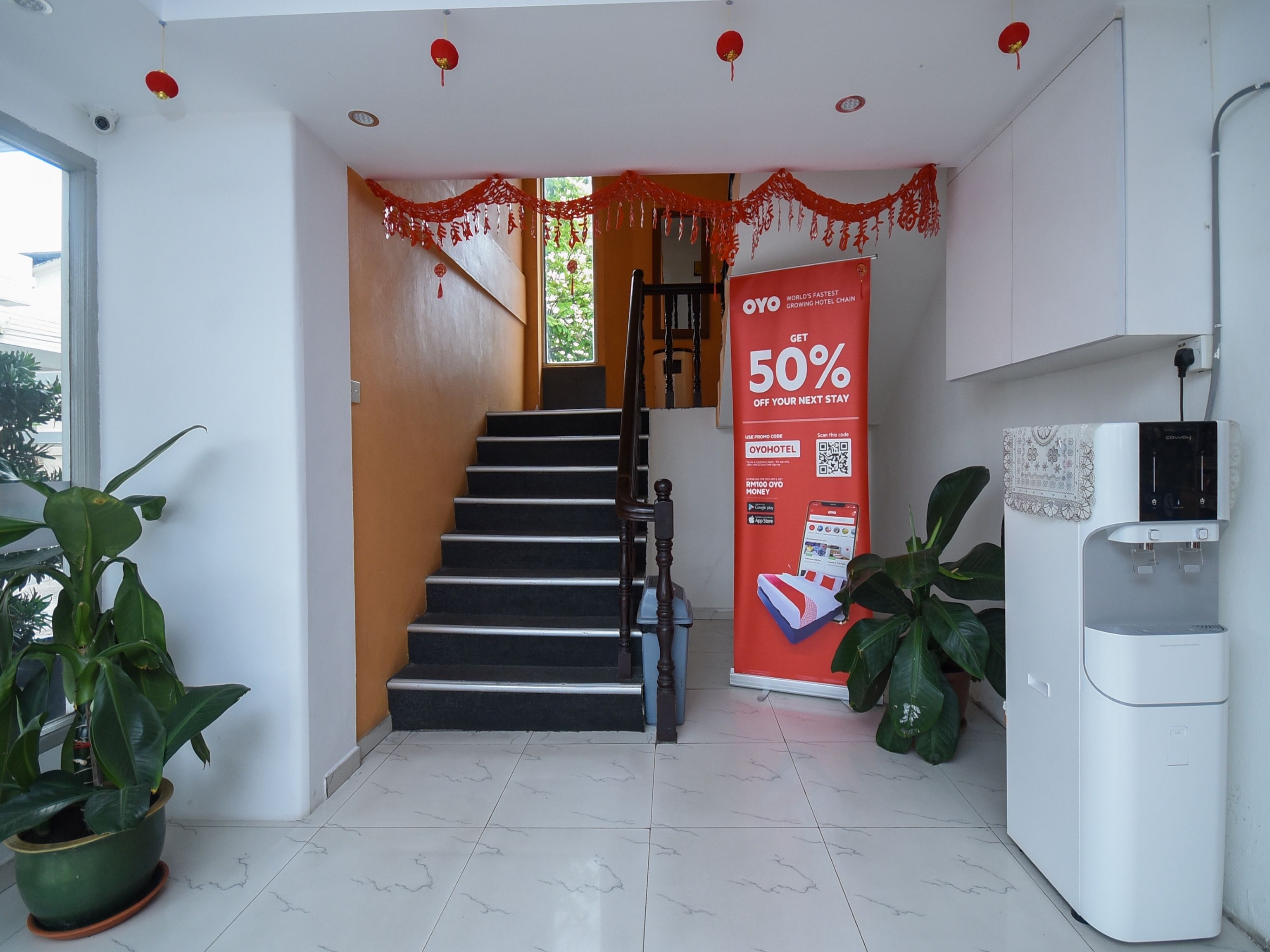 A hotel reception area with a staircase, a promotional banner, a water dispenser, and potted plants.