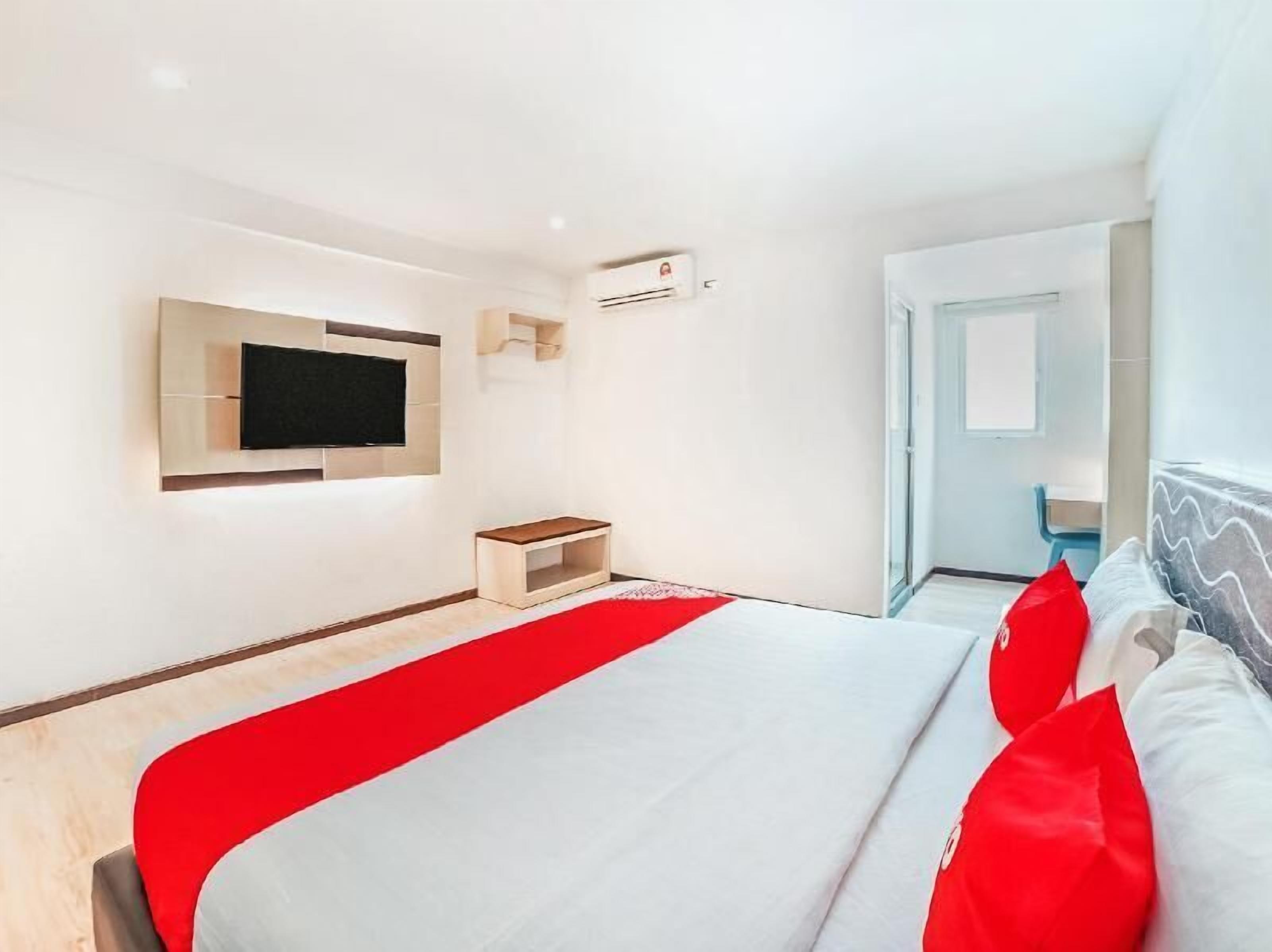 A modern hotel room with a large bed, a TV mounted on the wall, a small wooden bench, and a door leading to another room.