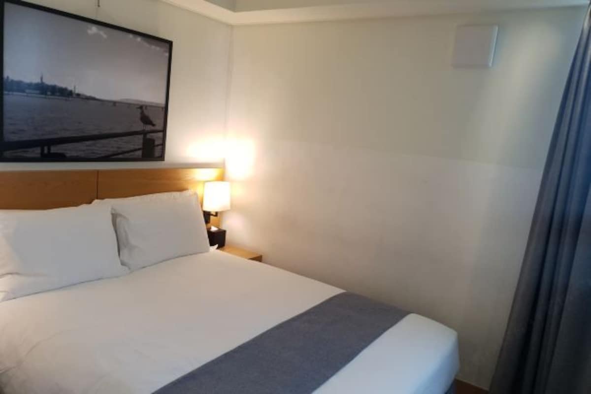 A hotel room with a large bed, a nightstand with a lamp, a framed picture on the wall, and a window with curtains.