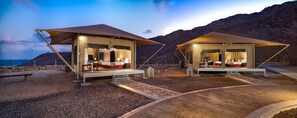 Two tents with beds and seating areas, set on a rocky beach with a mountainous backdrop.