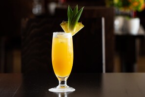 A tall glass with a yellow beverage and a pineapple garnish on a dark wooden table.