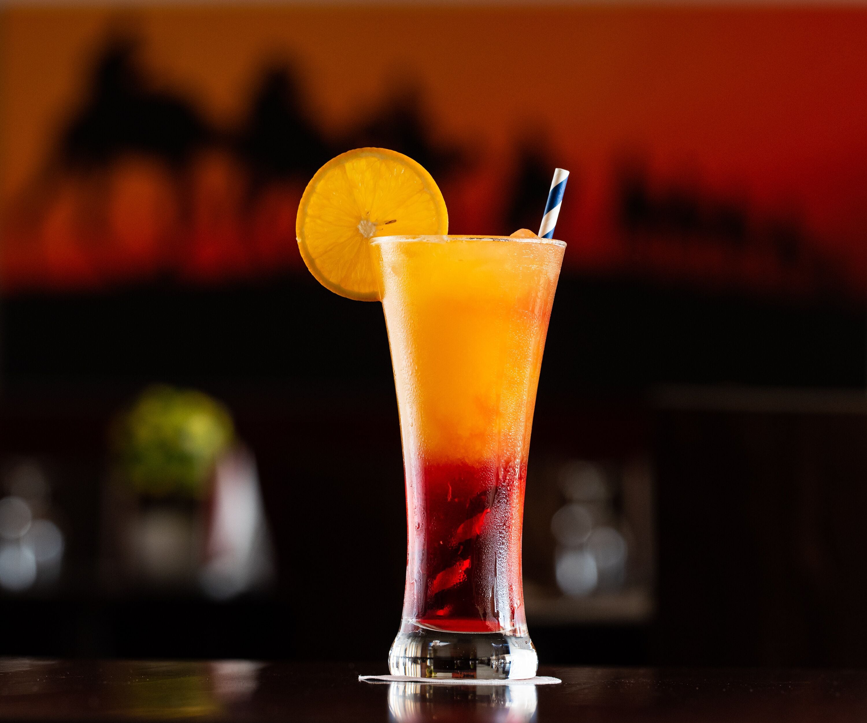 A tall glass with a gradient of red and orange liquid, garnished with a slice of orange and a striped straw.