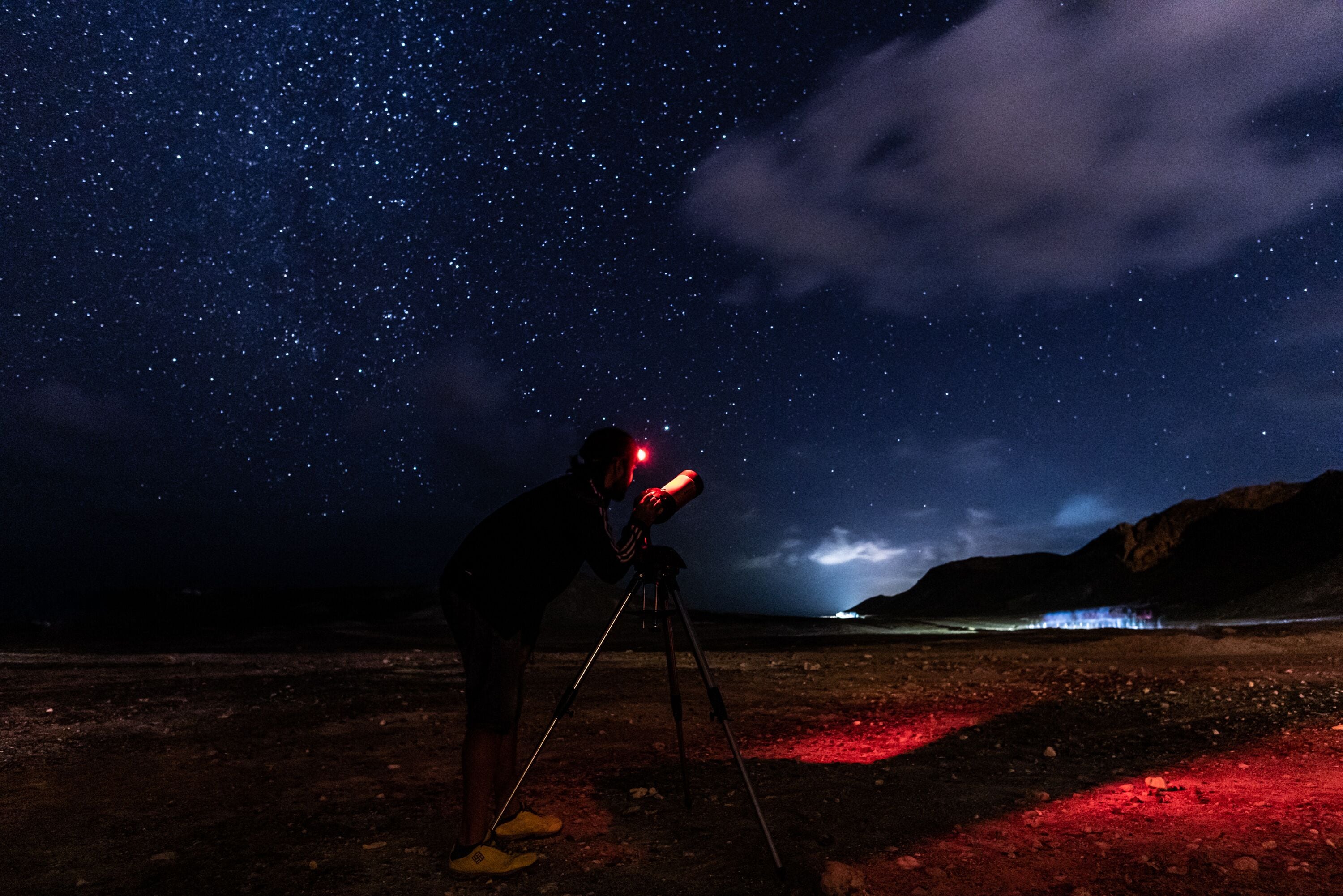 A person using a telescope on a tripod in a desert landscape at night.