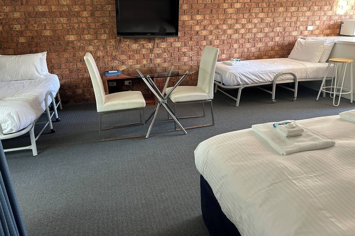 Quarto de hotel com parede de tijolos, televisão de tela plana, mesa de vidro com cadeiras, cama e um banquinho pequeno.