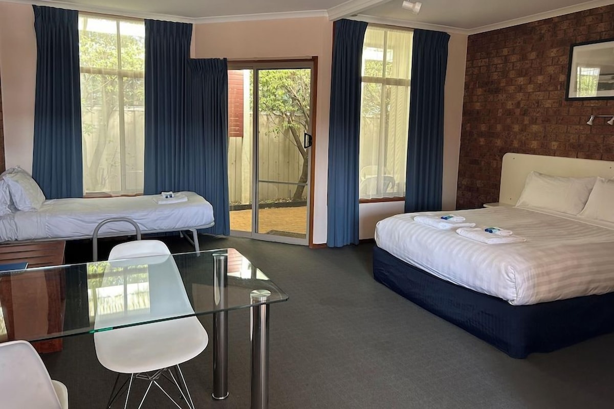 A hotel room with two beds, a glass table, and a view of the outdoors.
