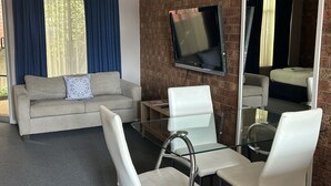 A modern living room with a sofa, two white chairs, a glass coffee table, and a flat-screen TV mounted on a brick wall. - Portarlington Beach Motel (Portarlington)