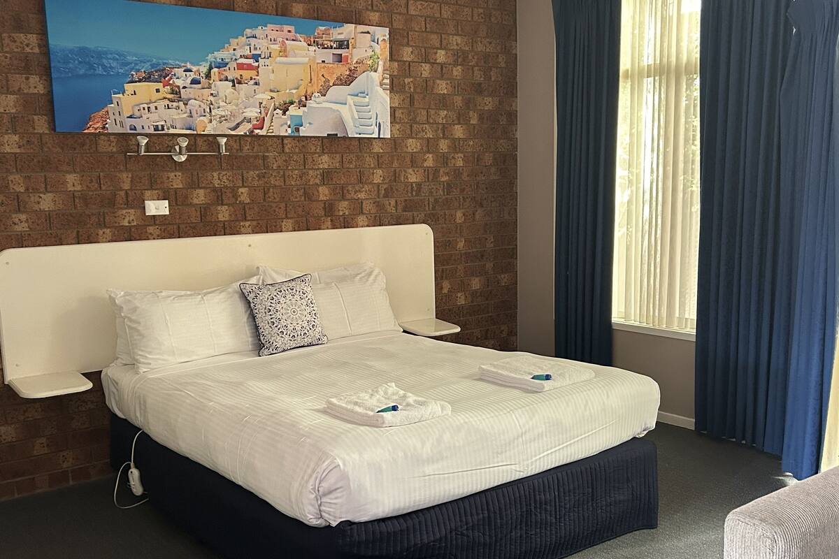 Quarto de hotel com uma cama grande, parede de tijolos, pintura de uma cidade costeira e uma janela com cortinas.