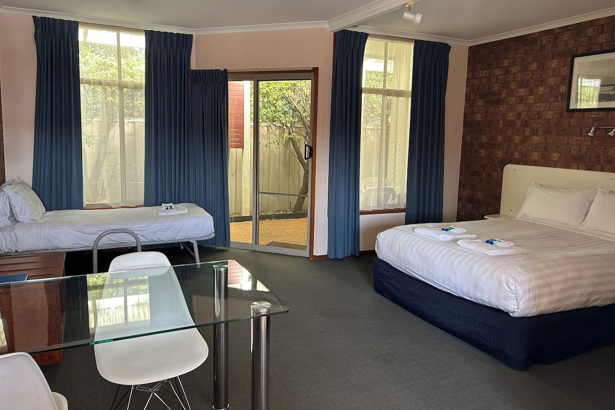 Quarto de hotel com duas camas de solteiro, uma mesa de vidro com cadeiras, uma escrivaninha de madeira e uma parede de tijolos.