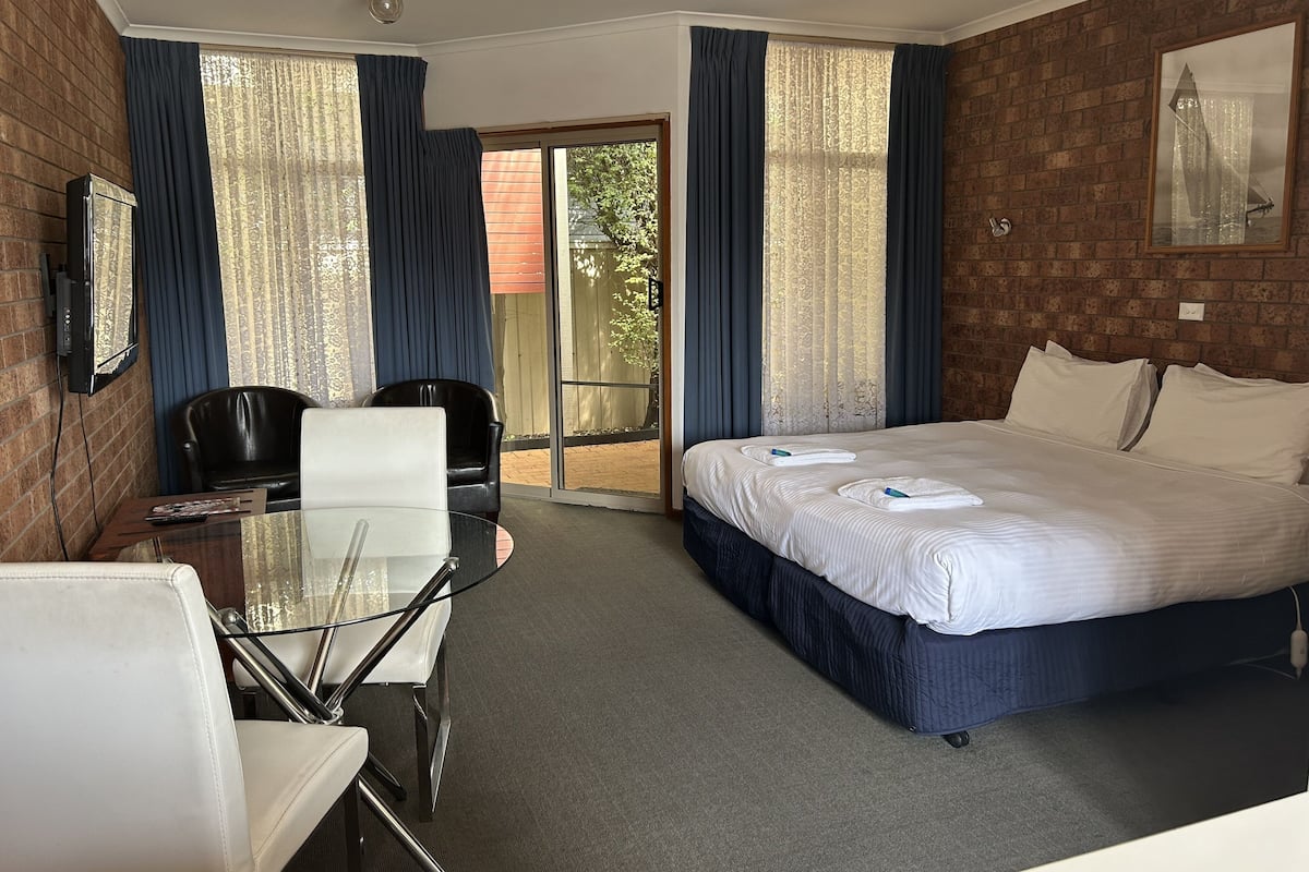 Quarto de hotel com uma cama, uma escrivaninha com cadeira, televisão presa à parede e uma porta de vidro deslizante para área externa.