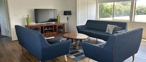 Un salon avec un canapé bleu foncé, deux fauteuils assortis, une table basse en bois, un téléviseur posé sur un meuble en bois et une grande fenêtre donnant sur un paysage verdoyant.