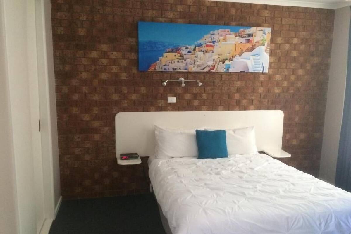 Quarto de hotel com parede em padrão de tijolos, cama branca com travesseiro azul, mesa de cabeceira com relógio e pintura de uma cidade costeira.