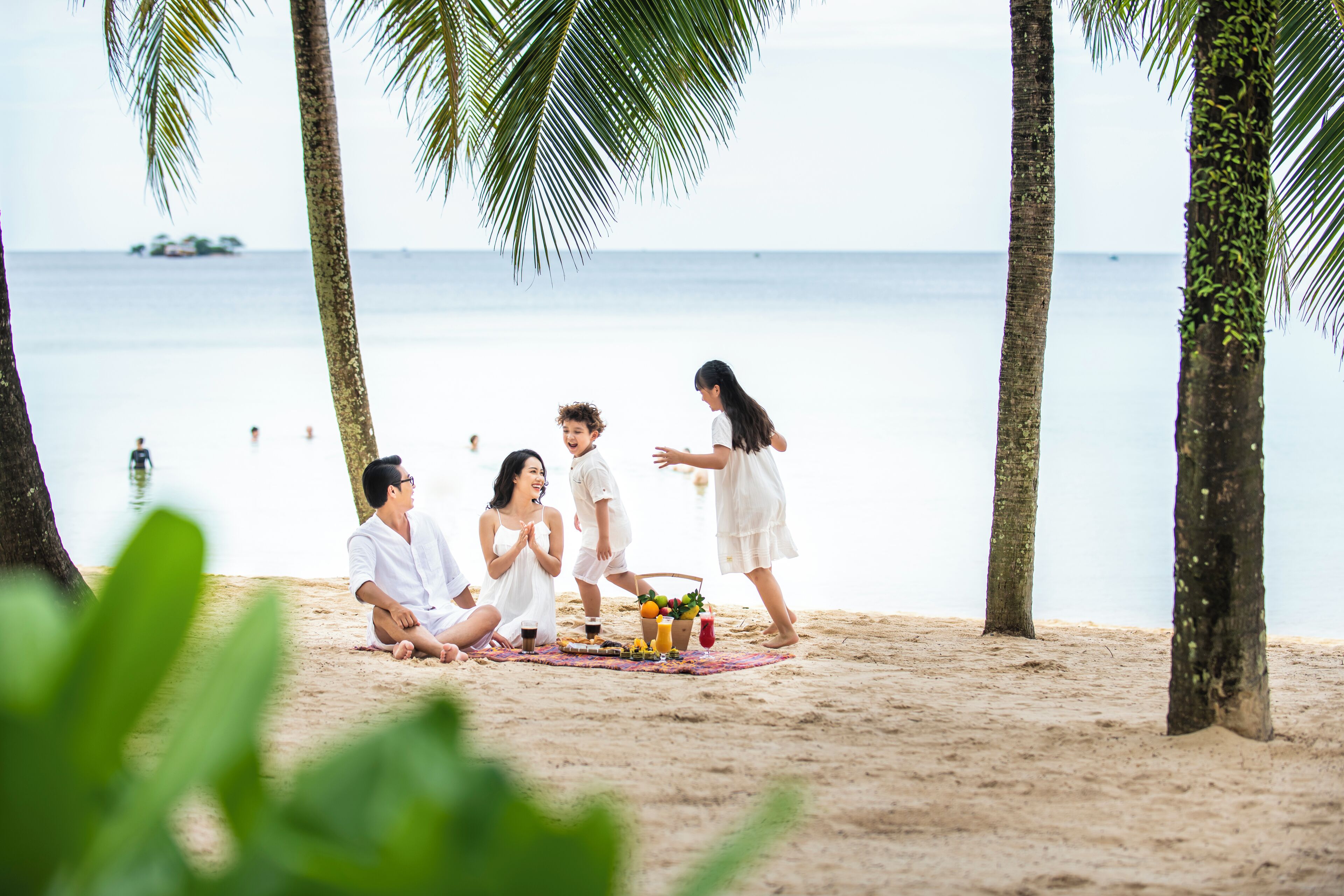 A group of people sitting on a beach with a picnic setup, palm trees, and the ocean in the background.