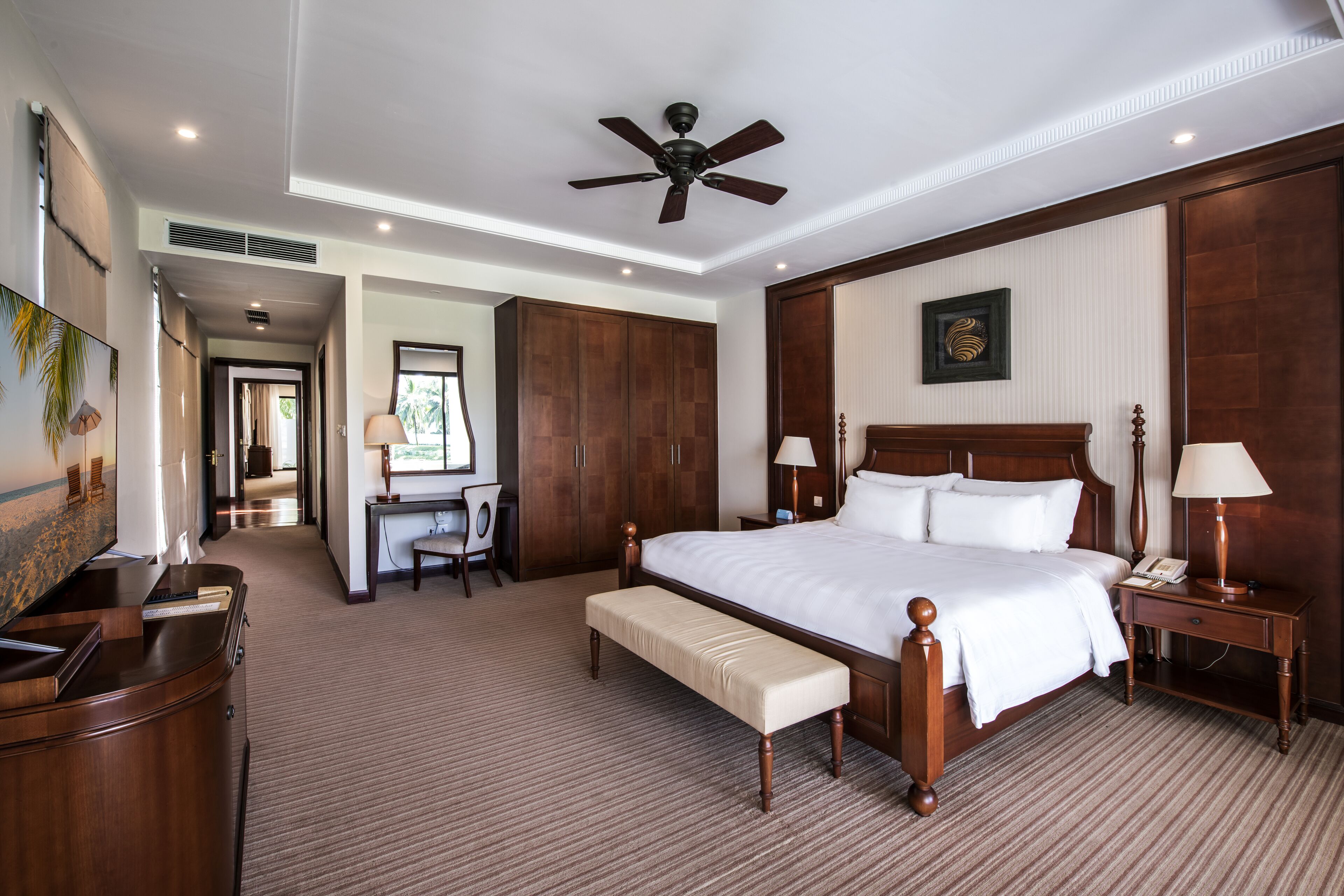 A spacious hotel room with a large bed, a desk, a chair, a bench, and a ceiling fan.