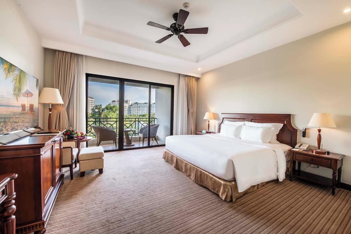 A hotel room with a large bed, a desk, a chair, a balcony with a view, and a ceiling fan.